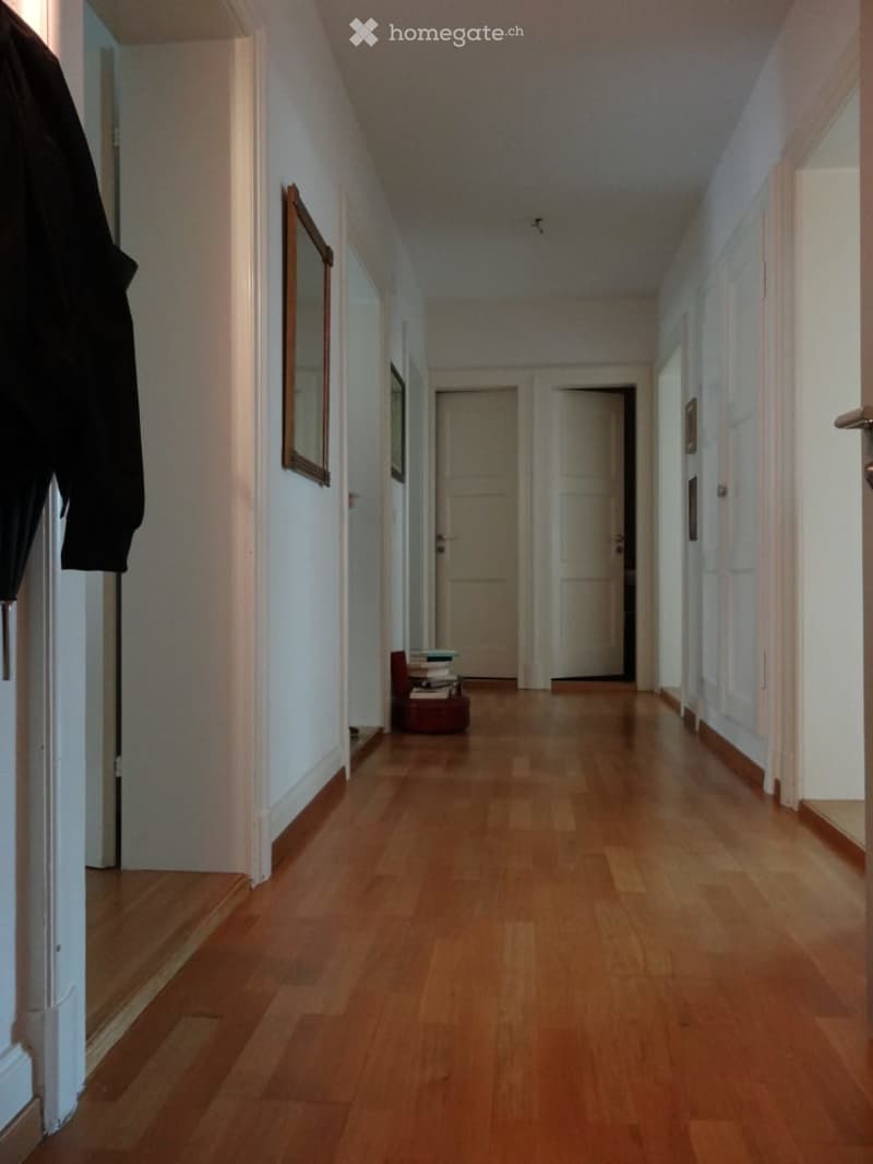 A long, narrow hallway with wooden flooring, white walls, and several doors leading to other rooms. A small stack of books and a mirror are visible on the left side.