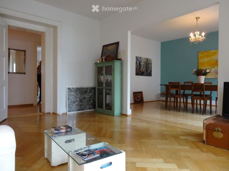 Spacious living and dining area with wooden flooring, white walls, and a blue accent wall. Features a glass coffee table, a green cabinet, a dining table with chairs, and a chandelier.