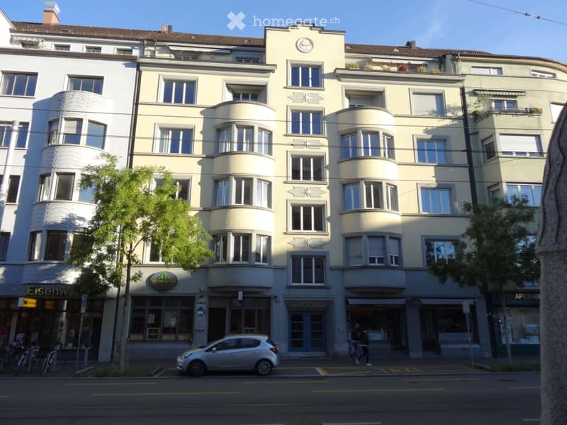 Multi-story residential building with cream-colored facade, large windows, and decorative architectural details. Ground floor includes commercial storefronts with signage.