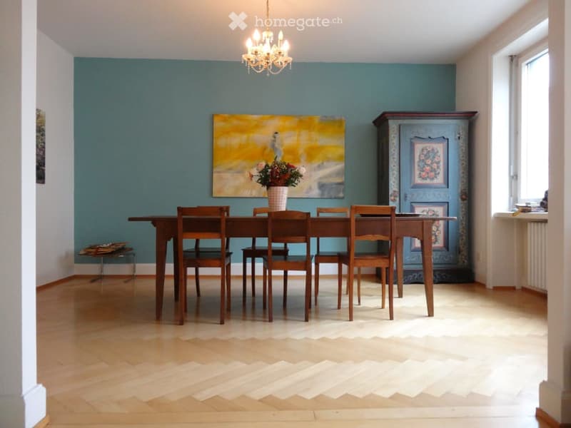Dining room with wooden floor, dining table, six chairs, flower vase, cabinet, painting, and chandelier.