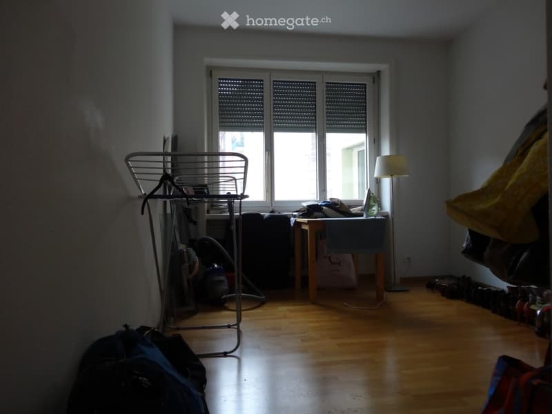 A small, furnished room with wooden flooring, a table with a lamp, a laptop, a coat rack, and a backpack on the floor.
