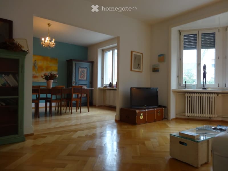 Open-plan living and dining area with wooden flooring, large windows, a dining table with chairs, a television on a trunk, a bookshelf, and a chandelier.