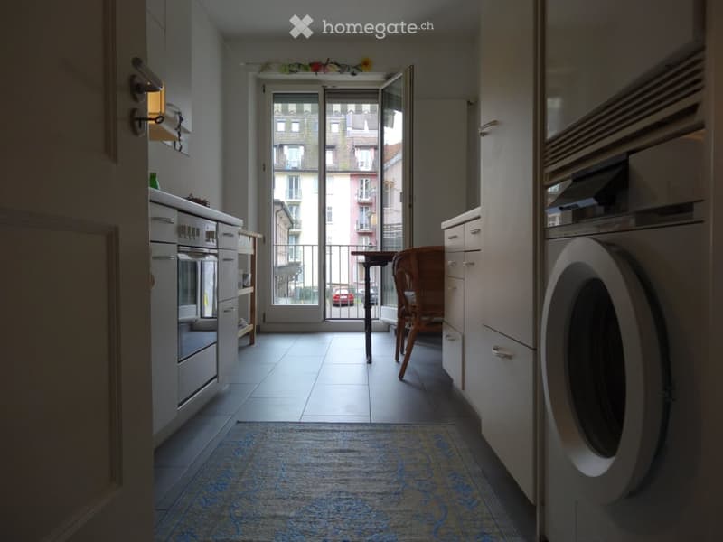 A modern kitchen with white cabinets, an oven, refrigerator, and washing machine. A small table and chair are near the balcony door, which offers a view of the city.