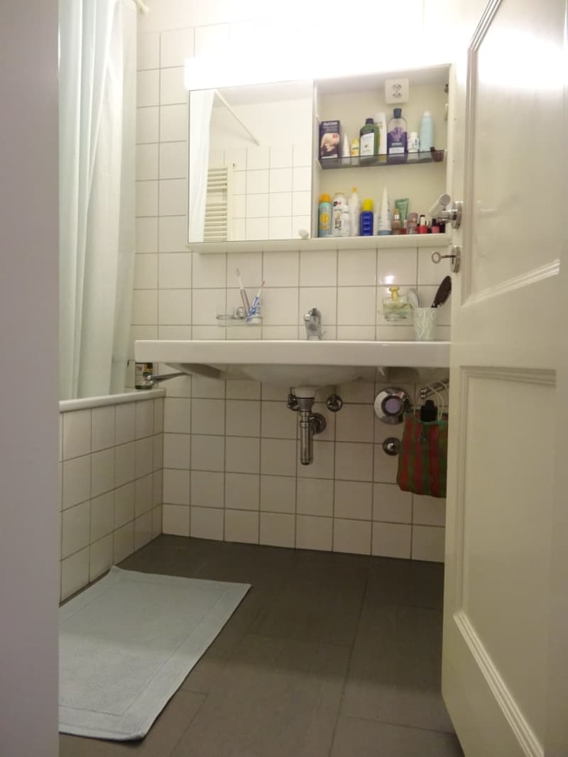 A small bathroom with a white tiled wall, a sink with a mirror above it, a toothbrush holder, a shelf with toiletries, and a towel on the floor.