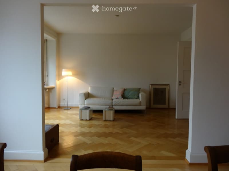 Bright living room with herringbone wooden flooring, white walls, a grey sofa with colorful cushions, a glass coffee table, and a floor lamp.