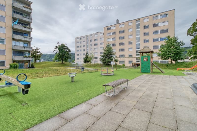 A well-maintained playground area with artificial grass, concrete pathways, and various play equipment including a seesaw, spring rider, climbing wall, and slide, surrounded by apartment buildings.