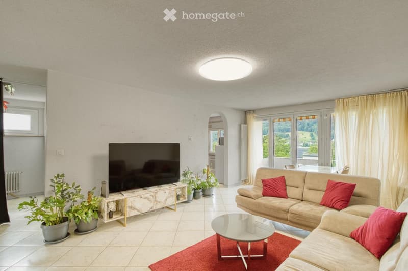 Modern living room with a beige sofa, red cushions, a glass coffee table, a large flat-screen TV, and several potted plants. The room features large windows with yellow curtains and a red area rug.