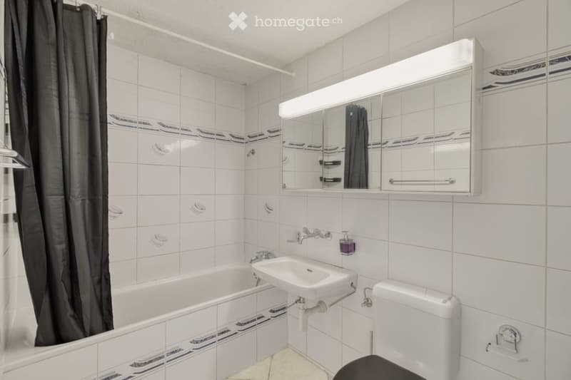 Modern bathroom with white tiles, bathtub, sink, toilet, mirror, and cabinet.