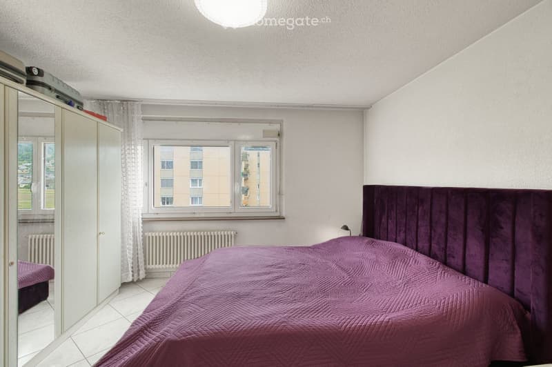 Bedroom with a large bed covered in a purple quilt, a matching headboard, a wardrobe, and a window with curtains overlooking a building.