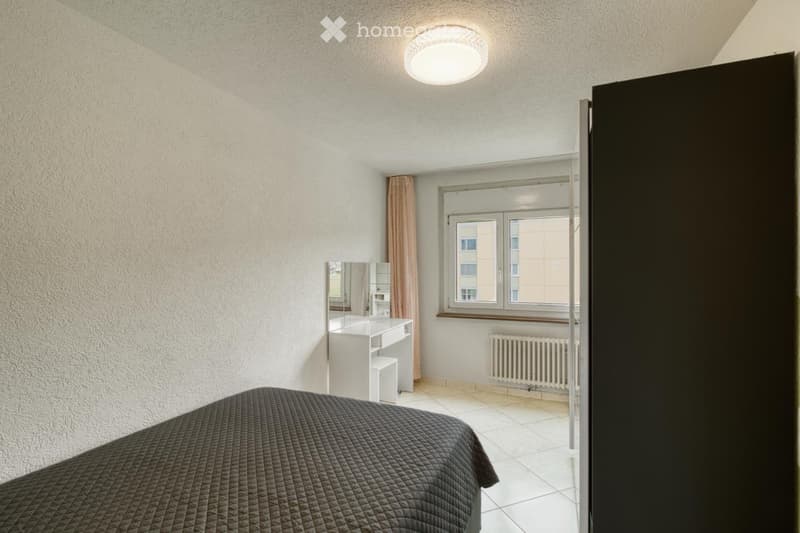 Modern bedroom with a dark quilted bed, white desk with mirror, radiator, and large window with curtains.