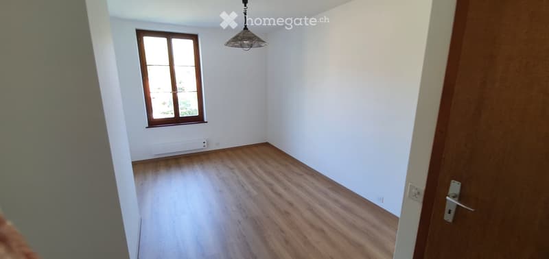 Empty room with wooden flooring, white walls, a single window with wooden frame, and a hanging light fixture.