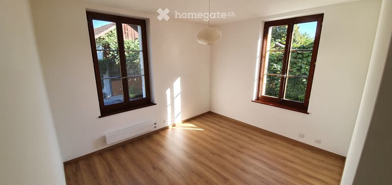 Empty room with wooden flooring, white walls, and large windows with wooden frames. Natural light streams in, and there is a hanging pendant light fixture.