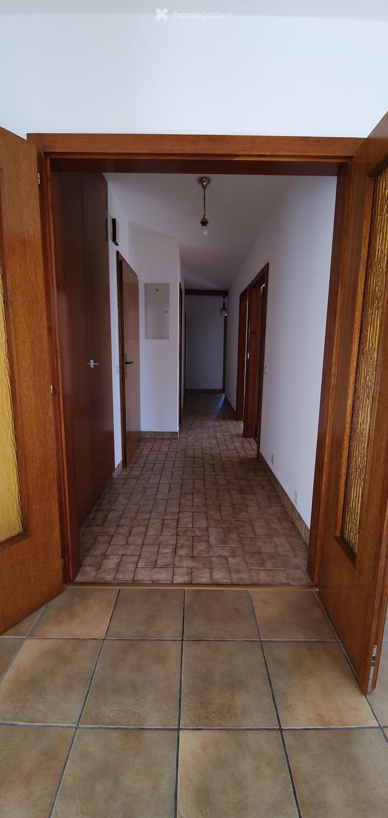 Empty hallway with tiled floor and wooden doors, featuring a ceiling light fixture and a visible electrical outlet on the wall.