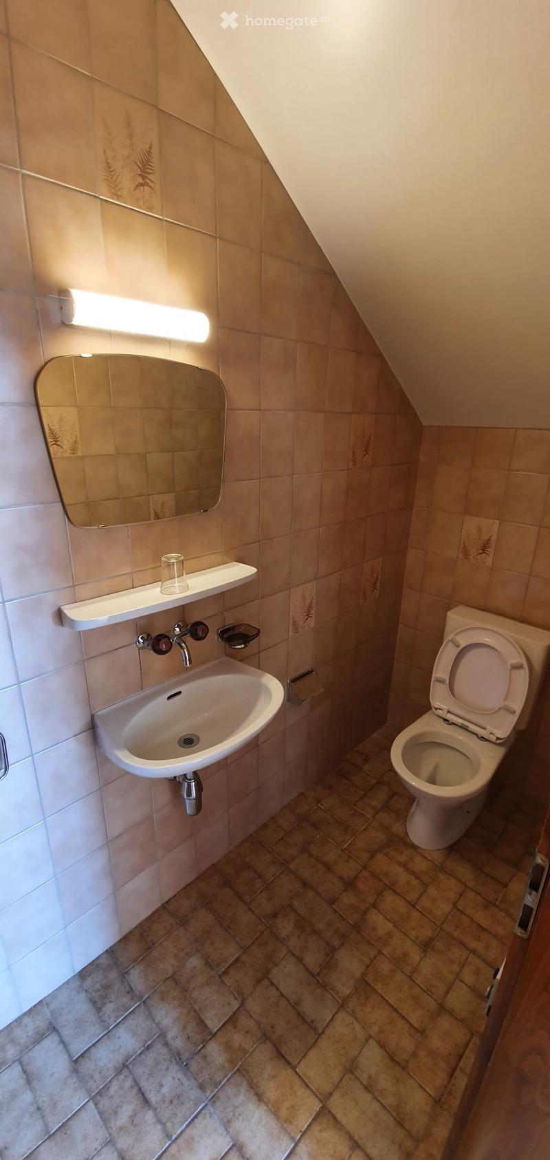 Small bathroom with beige tiles, a sink with a mirror above it, a toilet, and a small shelf holding a glass.