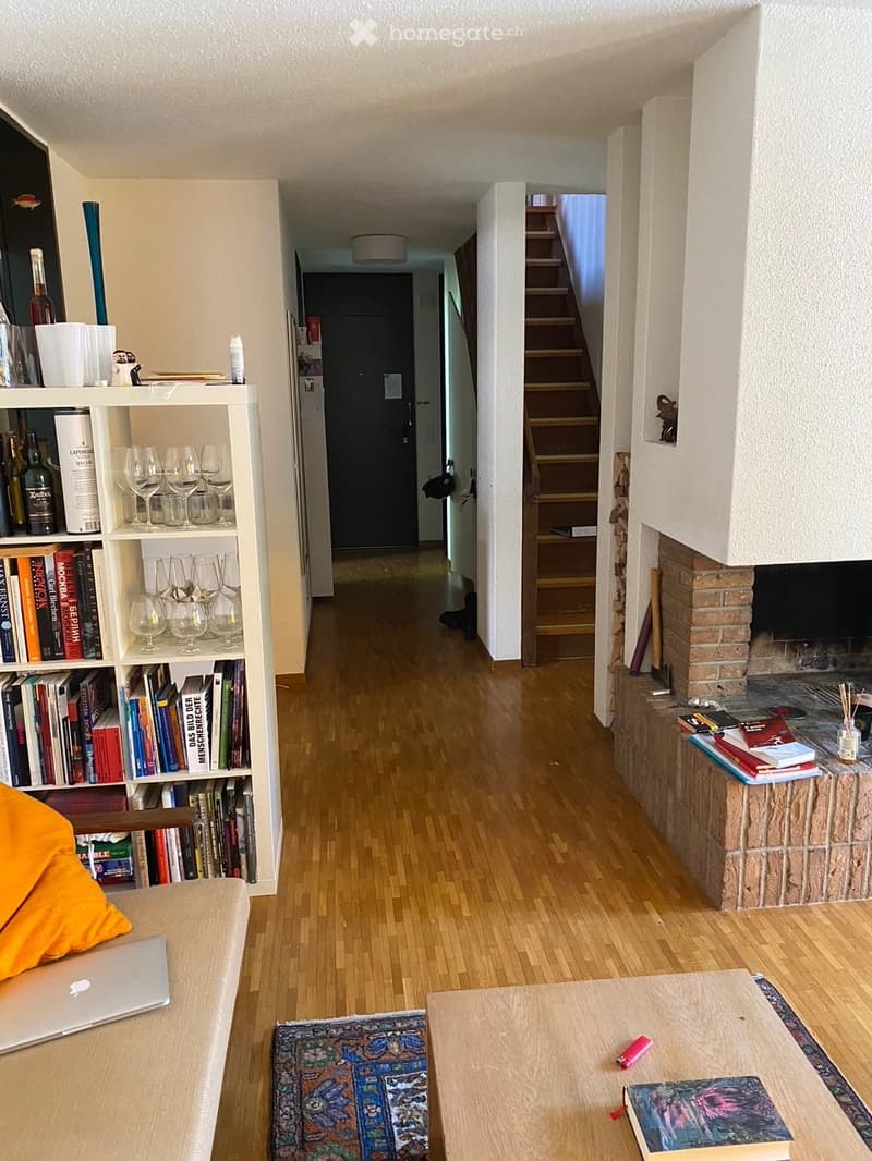 Modern living room with a fireplace, bookshelf filled with books and wine bottles, a laptop on a sofa, and a cozy atmosphere.