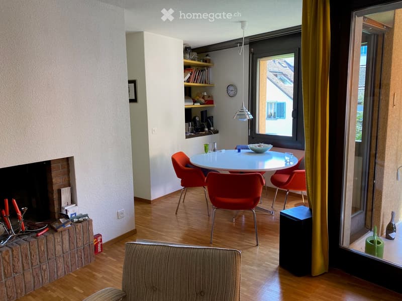 A modern, well-lit room with a hanging chair, a small table, shelves, a clock, and a tall lamp near the window.
