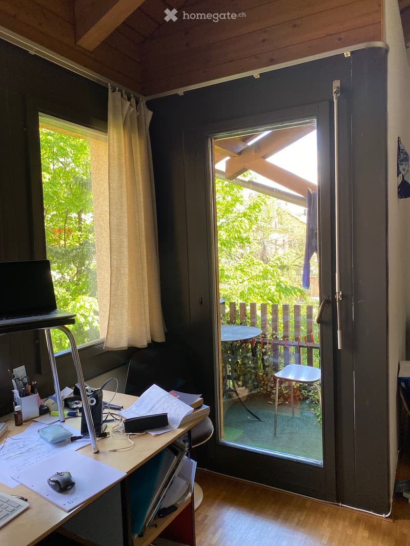 A well-lit room with a wooden desk, laptop, chair, and a large window with a curtain, overlooking a green outdoor area.
