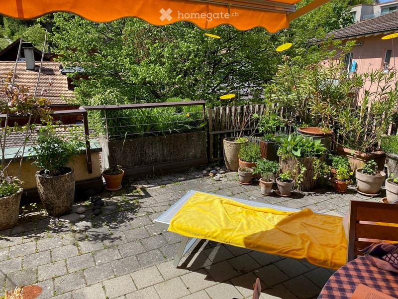A sunny outdoor patio with a lounge chair, potted plants, and a wooden fence, surrounded by greenery and neighboring houses.