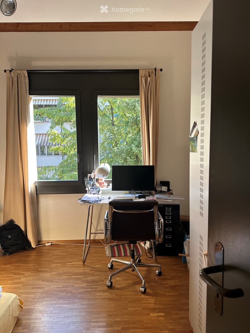A furnished room with a wooden floor, a large window with curtains, a desk with a chair, a screen, and a backpack on the floor.