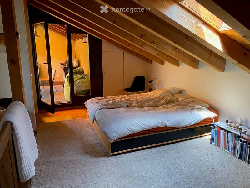 Cozy attic bedroom with wooden slanted ceilings, skylight windows, a bed with white bedding, a bookshelf, a chair, and a mirror. The room features a warm, inviting atmosphere with soft lighting and a carpeted floor.