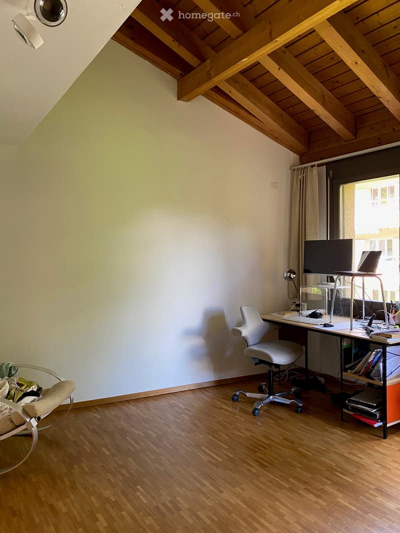A small, well-lit room with wooden flooring and a desk setup, including a chair, laptop, and screen. A shelf is visible on the left side.