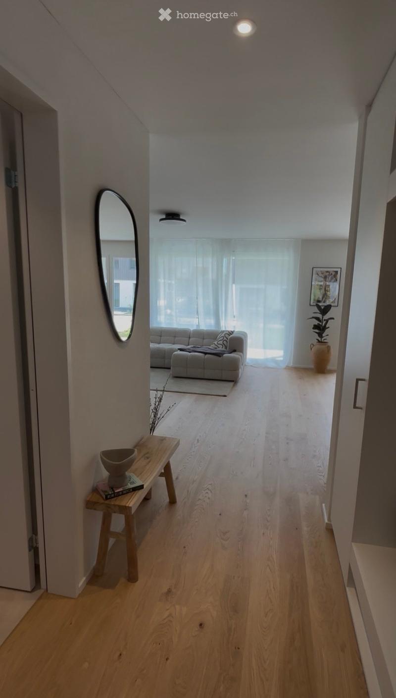 A modern hallway with light wooden flooring, white walls, and recessed lighting. Features include a small wooden bench with a bowl and books, a round mirror on the wall, and a view into a living room area with a grey sofa and a potted plant.