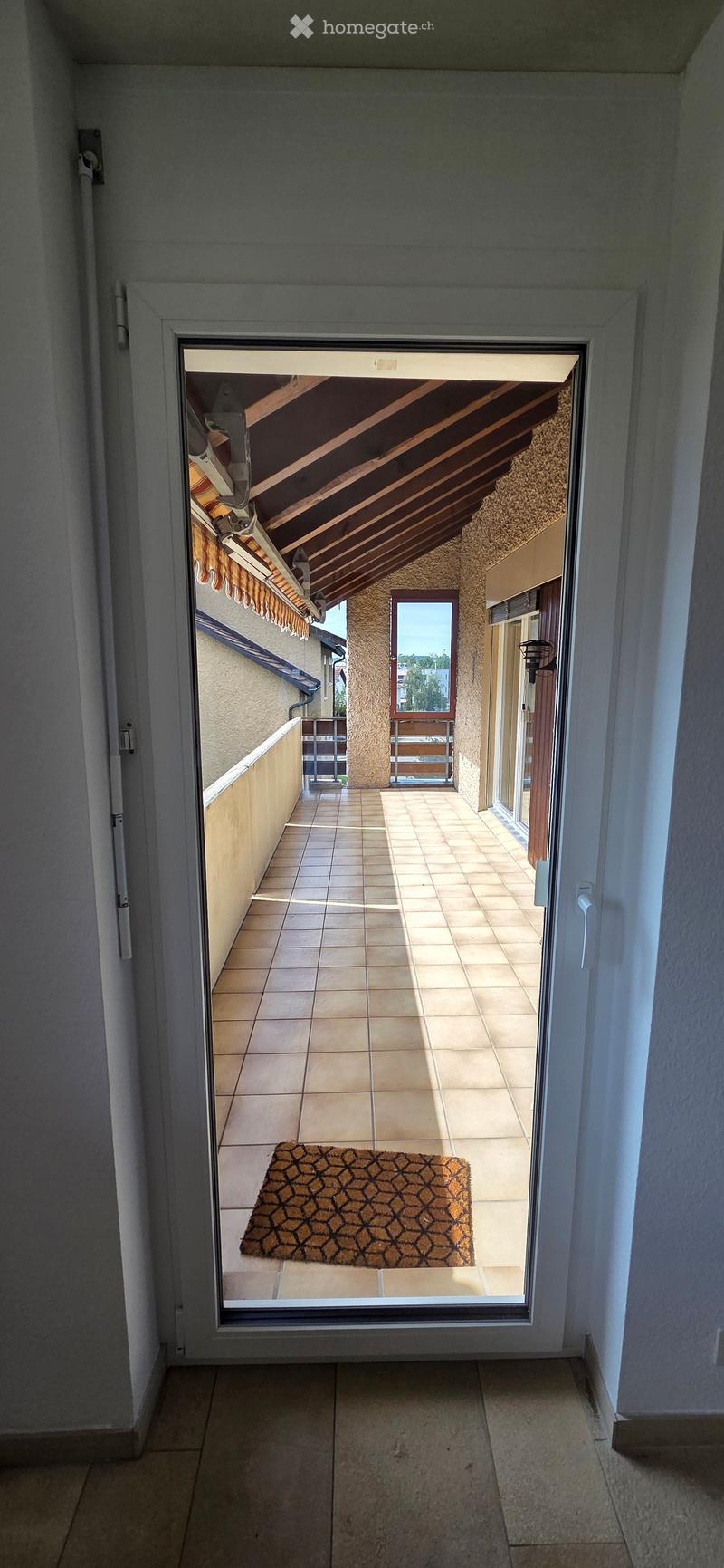 View through an open sliding door onto a tiled balcony with a patterned doormat, wooden railings, and a covered roof area.