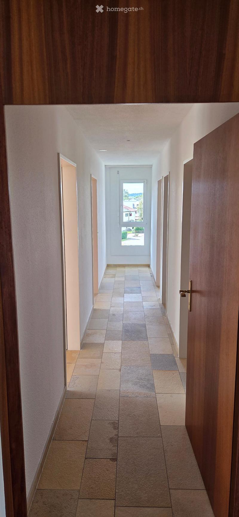 A long, narrow hallway with tiled flooring and light-colored walls, featuring several doors and a window at the far end that lets in natural light.