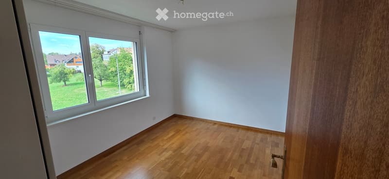 Empty room with wooden flooring, white walls, and large windows offering a view of a green area with trees and houses.