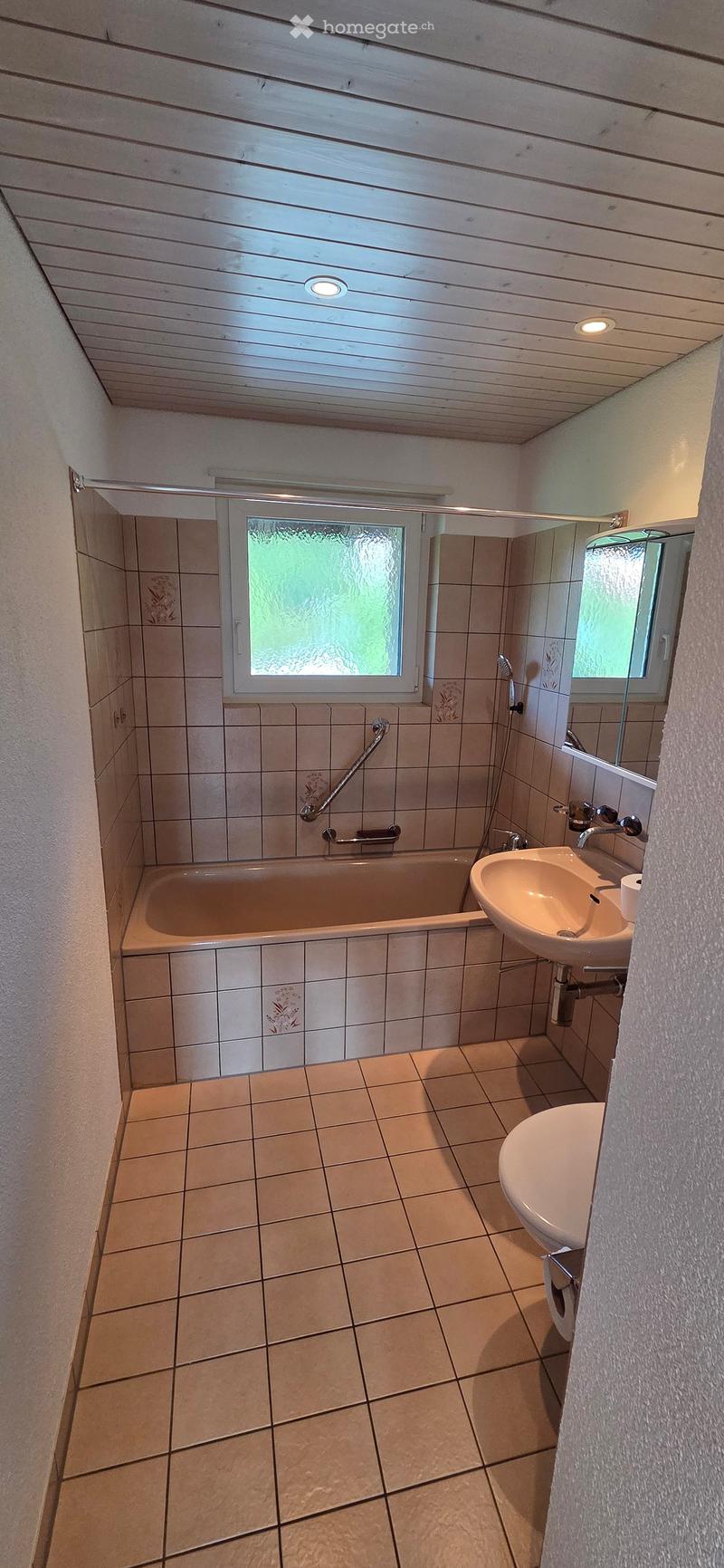Bathroom with tiled walls and floor, featuring a bathtub, sink, mirror, and toilet. The room has a window with frosted glass and built-in lighting.