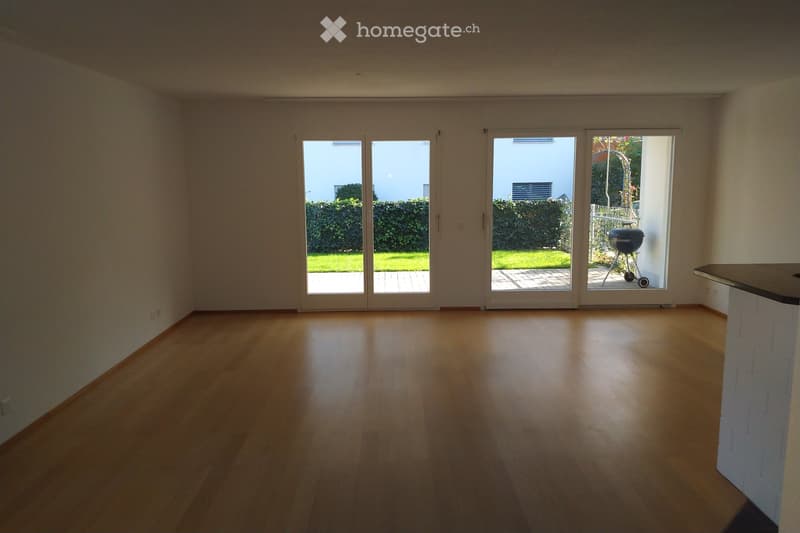Empty room with light wooden flooring, large sliding glass doors leading to a garden, and a white brick wall on one side.