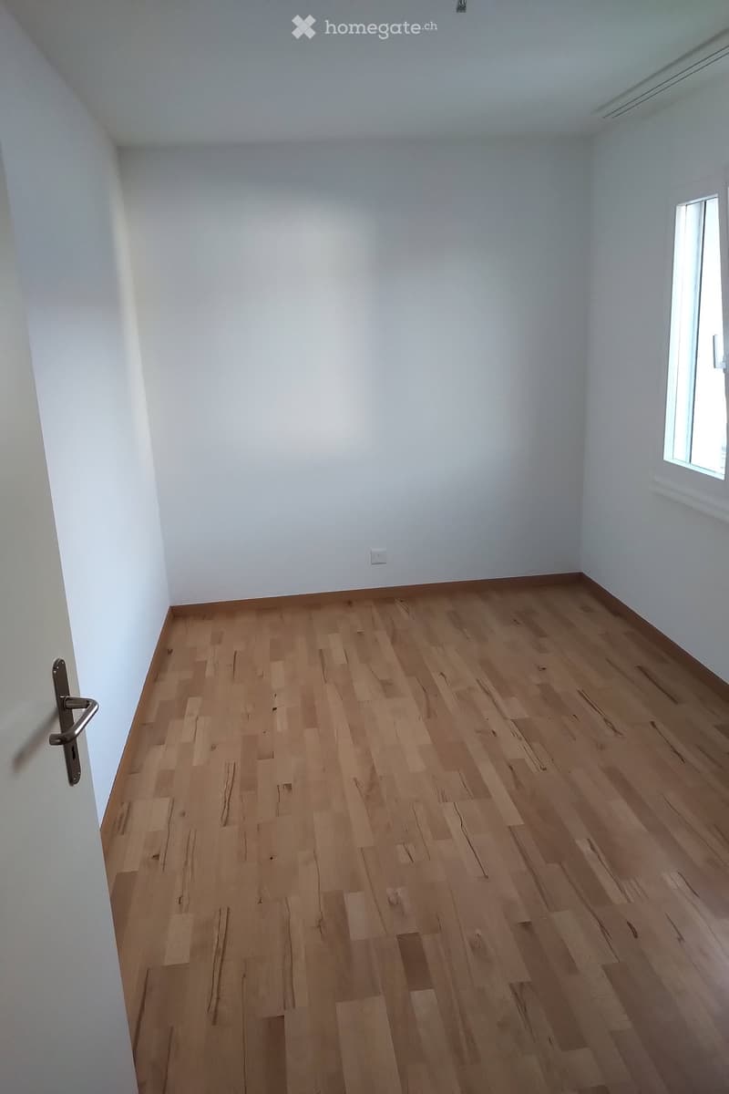 Empty room with light wooden flooring and white walls, featuring a single window and a door with a handle.