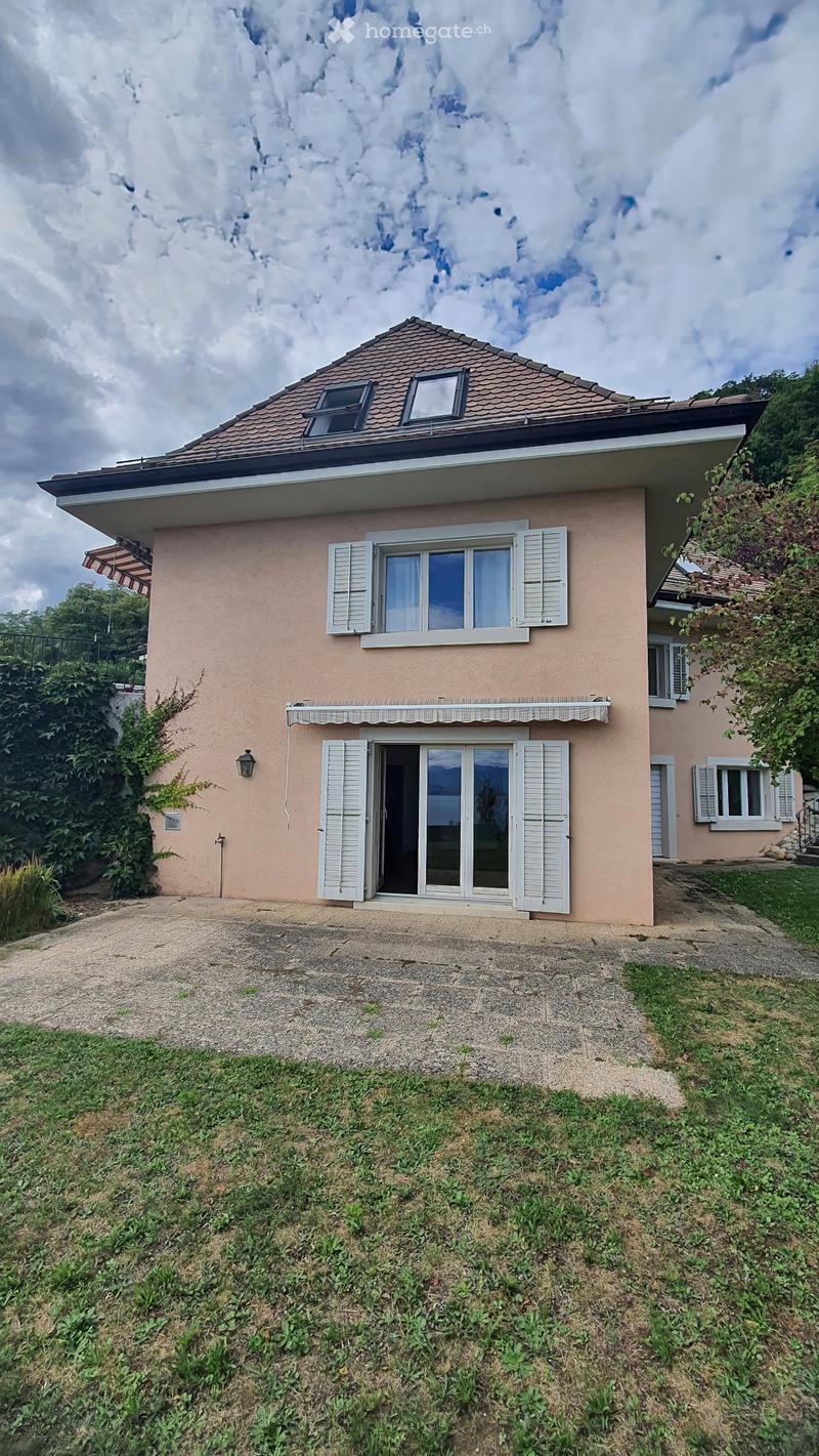 Two-story house with beige exterior, tiled roof, multiple windows with shutters, and a covered entrance.