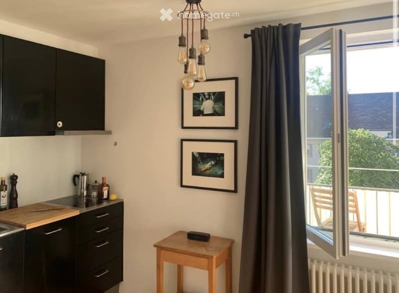 Modern kitchen with black cabinets, a wooden cutting board, a small wooden stool, and two picture frames on the wall. A window with a curtain opens to a balcony.