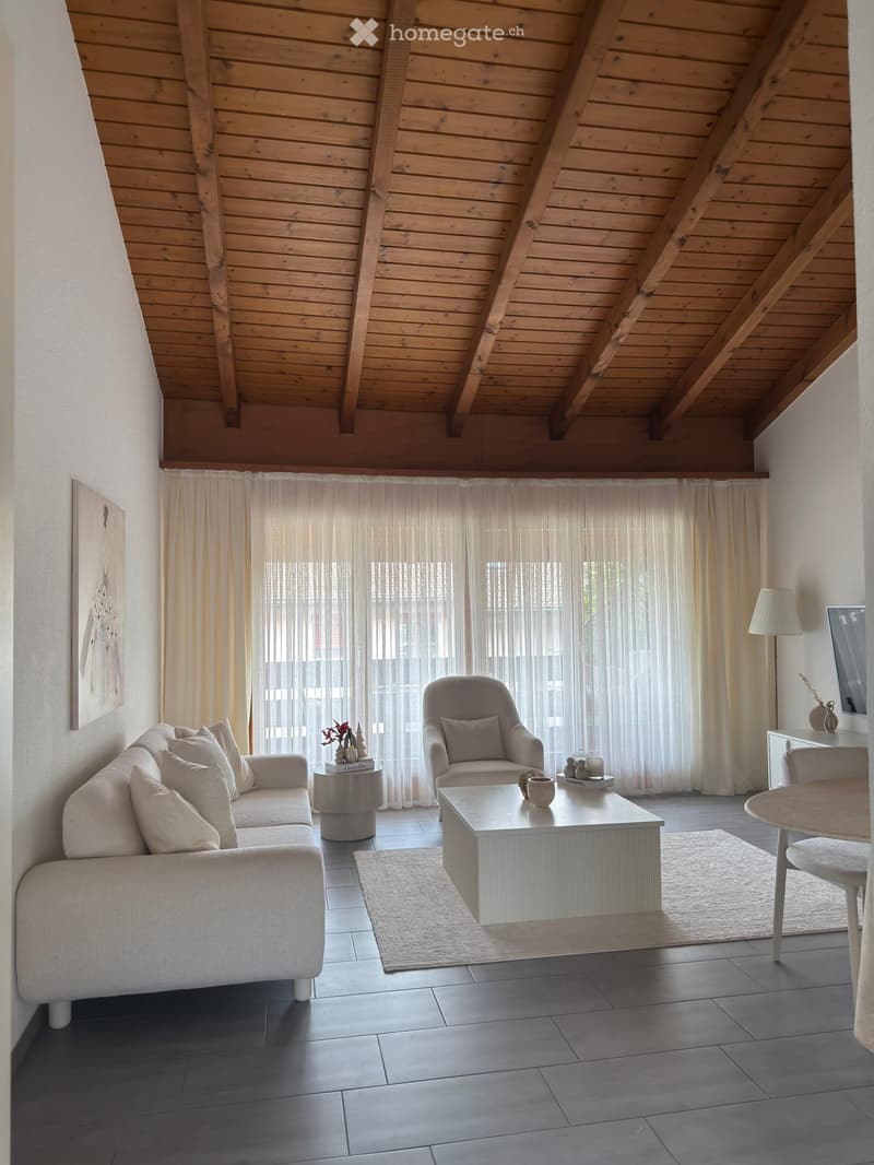 Modern living room with a grey sofa, armchair, coffee table, side table, and dining area with a table and chairs. The room features large windows with sheer curtains, a wooden slanted ceiling, and a neutral color palette.
