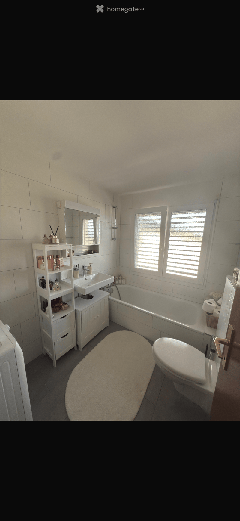 Modern bathroom with white tiles, a bathtub, toilet, sink, mirror, and a white bathroom cabinet with toiletries. The room features a large window with blinds and a plush beige rug on the floor.