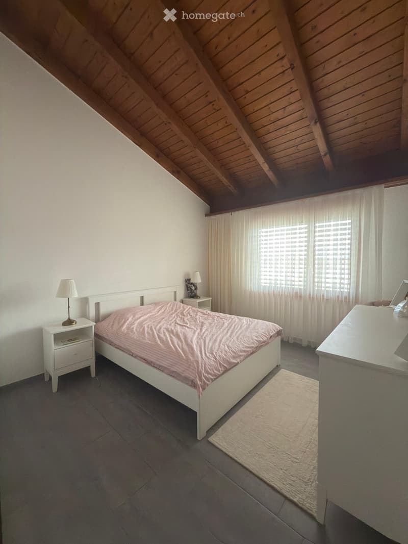 Cozy attic bedroom with wooden slanted ceilings, grey tiled floor, white walls, and large windows with white curtains. Features a white bed with pink bedding, two nightstands with lamps, a small rug, and a mirror on the dresser.