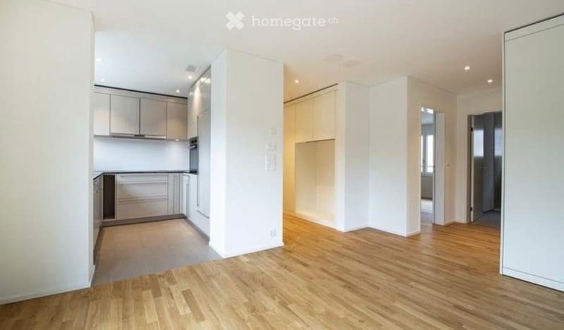 Open-plan unfurnished room with light wooden flooring, white walls, and a small kitchen area featuring white cabinets and stainless steel appliances.