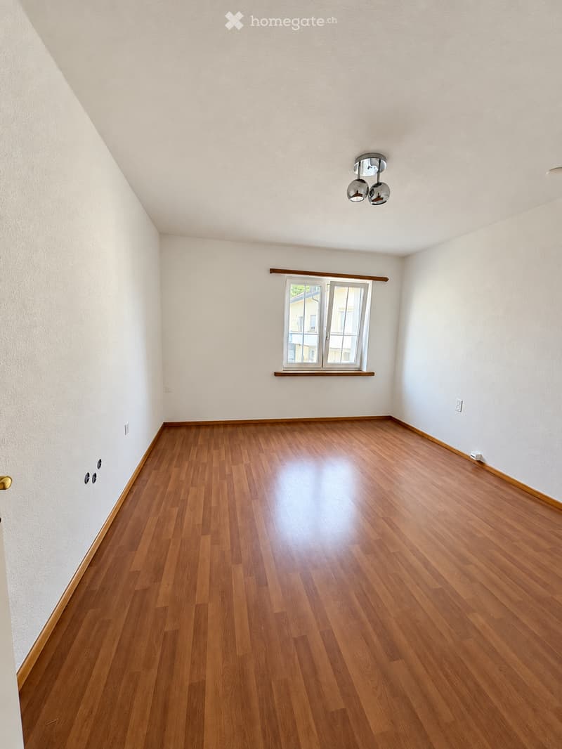 Empty room with wooden flooring, white walls, and a window with a wooden sill. The room features a modern ceiling light fixture and a door with a golden handle.