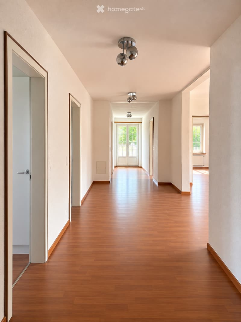 Spacious hallway with polished wooden flooring, light-colored walls, and modern ceiling lights. Multiple doors lead to adjacent rooms, with a visible window at the far end allowing natural light to enter.