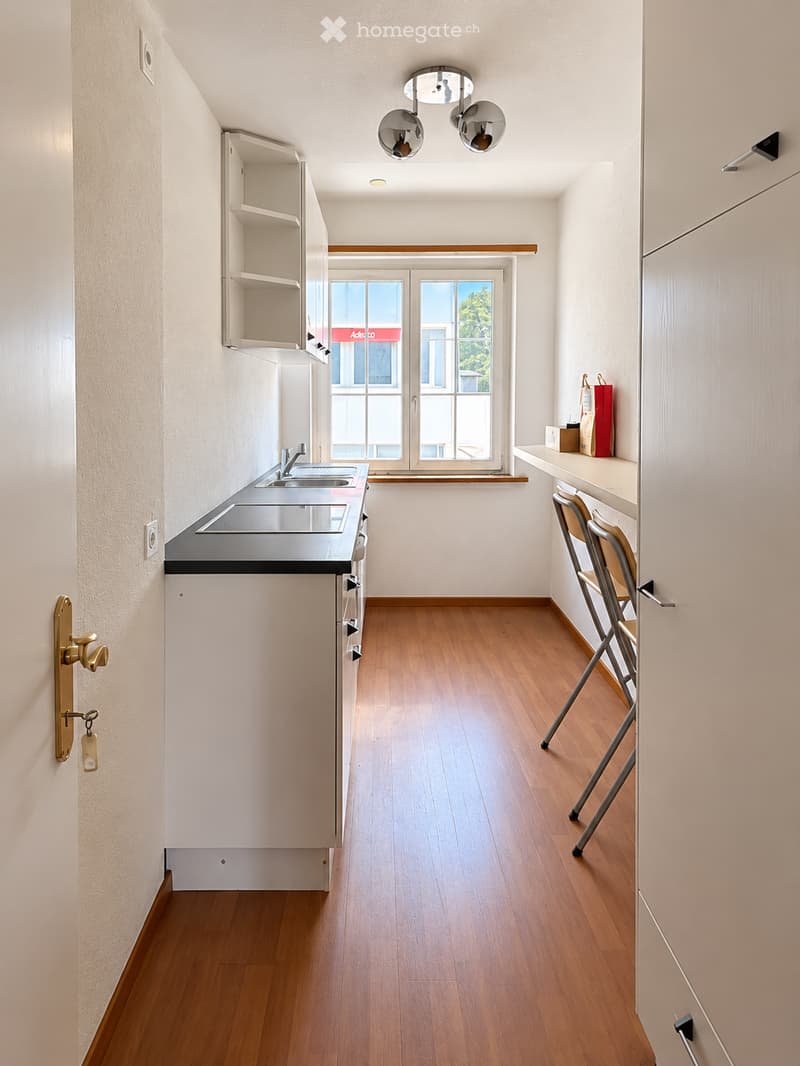 Empty kitchen with white cabinets, wooden floor, and a window with a view of greenery. No furniture or appliances are present.