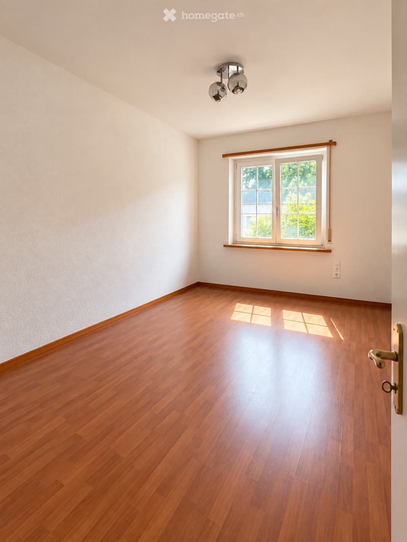 Empty room with wooden flooring, white walls, and a large window with a wooden frame. The room features a modern ceiling light fixture and a door with a brass handle.