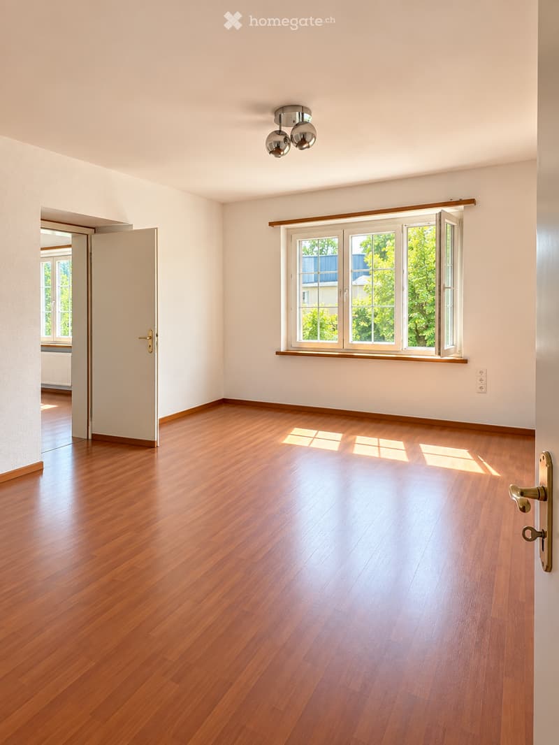 Empty room with wooden flooring, white walls, large window with wooden frame, and a ceiling light fixture.