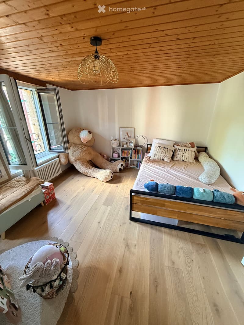 Cozy bedroom with wooden floors, slanted wooden ceiling, and large window. Features a bed with pillows, a large teddy bear, a bookshelf, and a basket with toys.
