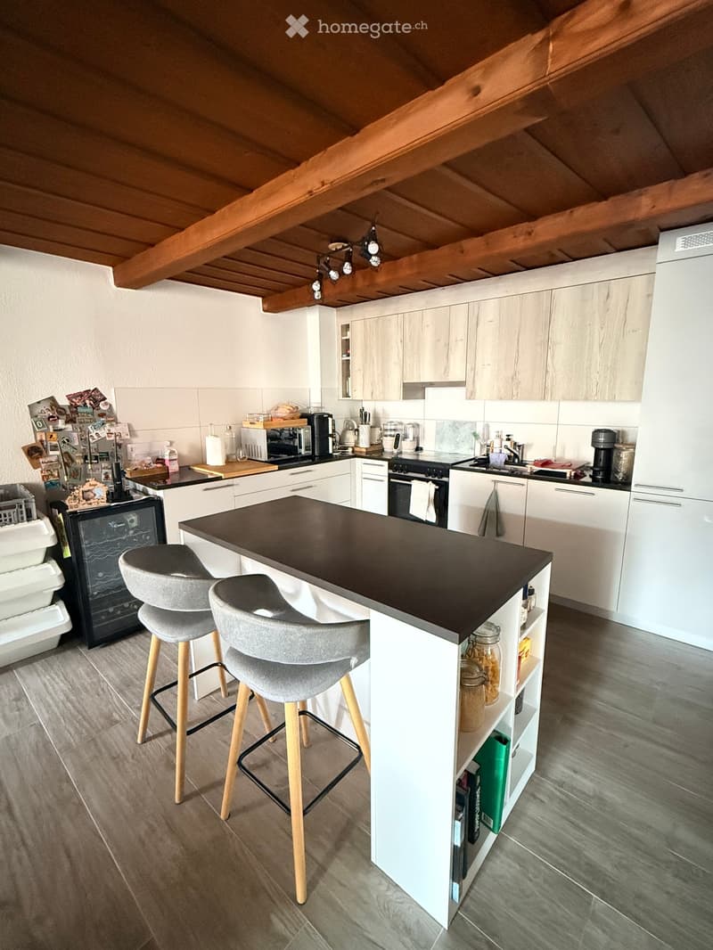 Modern kitchen with white cabinets, dark countertops, and a central island. Includes a stove, refrigerator, microwave, coffee maker, and two stools.