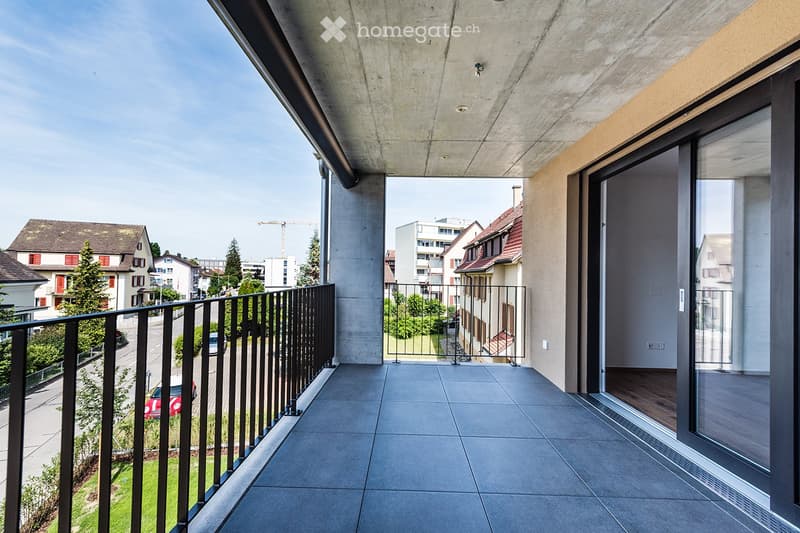 Modern balcony with grey tiled floor, black metal railing, and large glass doors leading to the interior. The view shows a residential street with houses, trees, and parked cars.