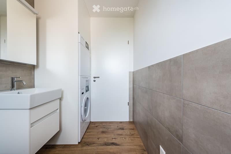 Modern bathroom with a white sink, chrome faucet, built-in washing machine, and light grey tiled wall.