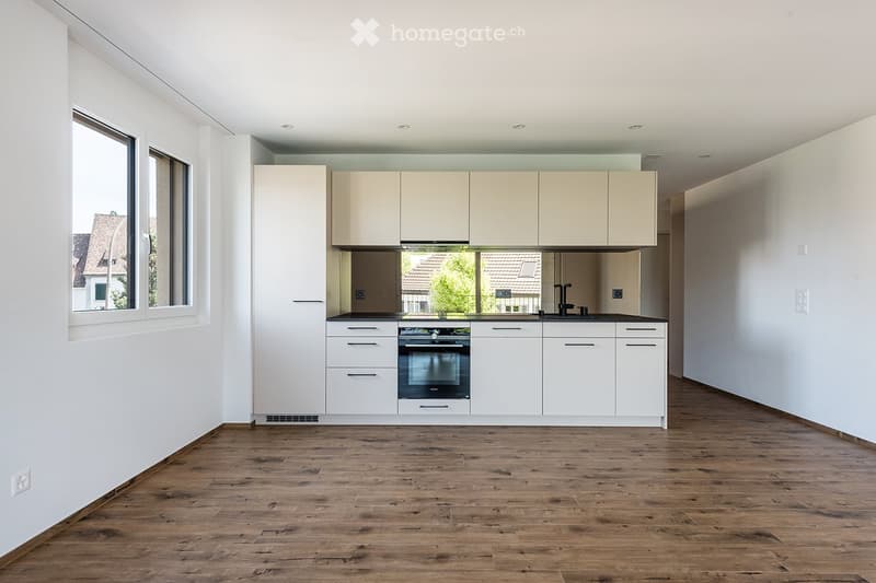 Modern kitchen with white cabinets, a built-in oven, and a sink, featuring wooden flooring and large windows.