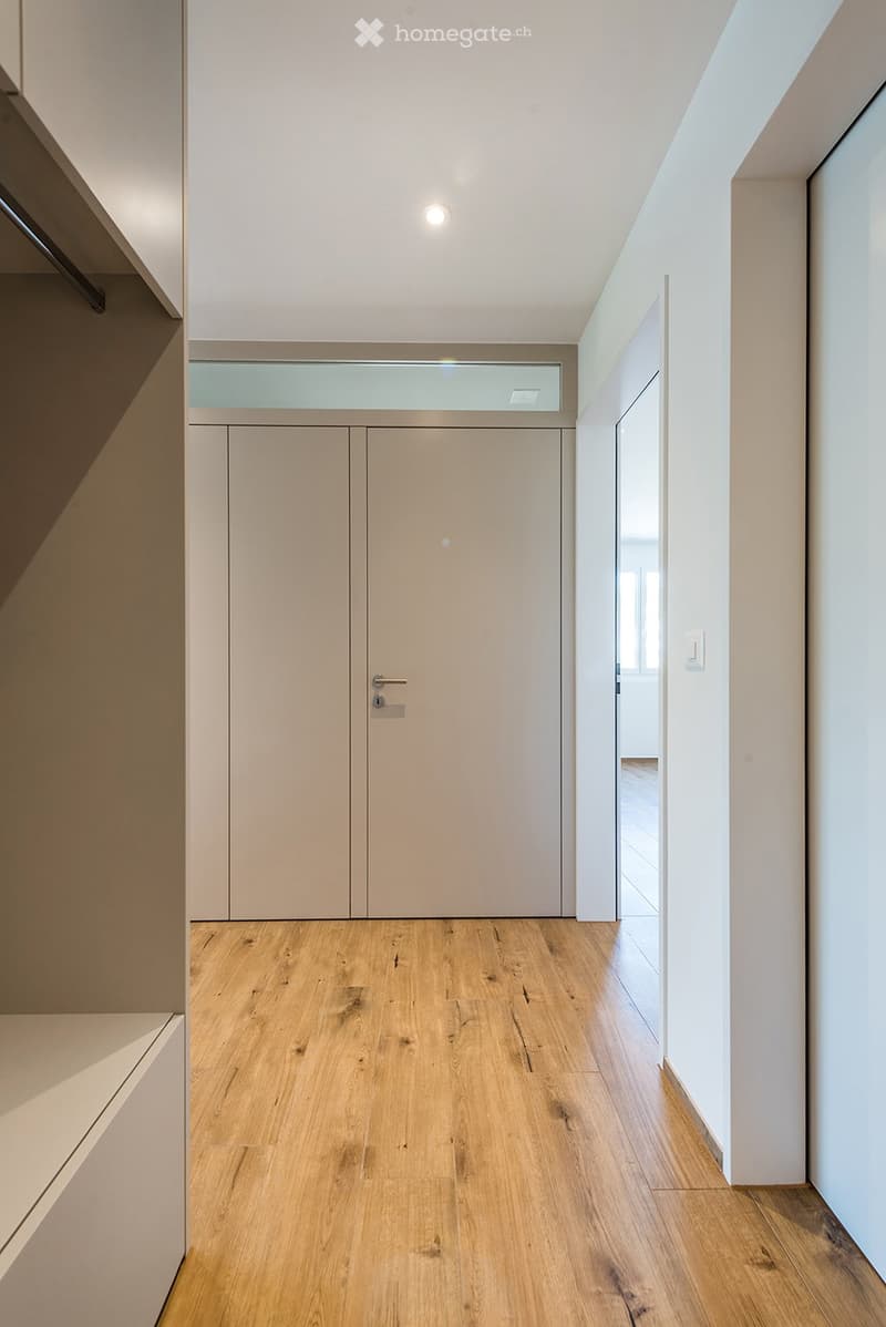 Modern hallway with light wooden flooring, white walls, and recessed lighting. Features a closed double door and an open doorway leading to another room.