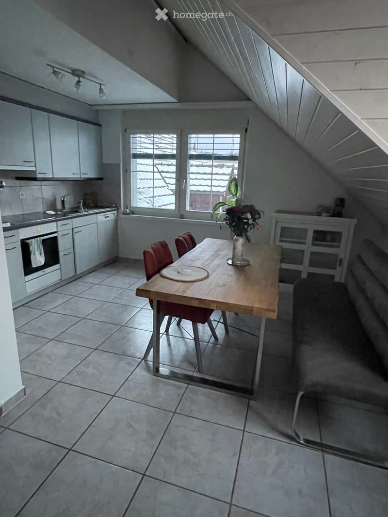 Attic kitchen with white cabinets, tiled floor, wooden dining table with chairs, and a vase with flowers on the table.